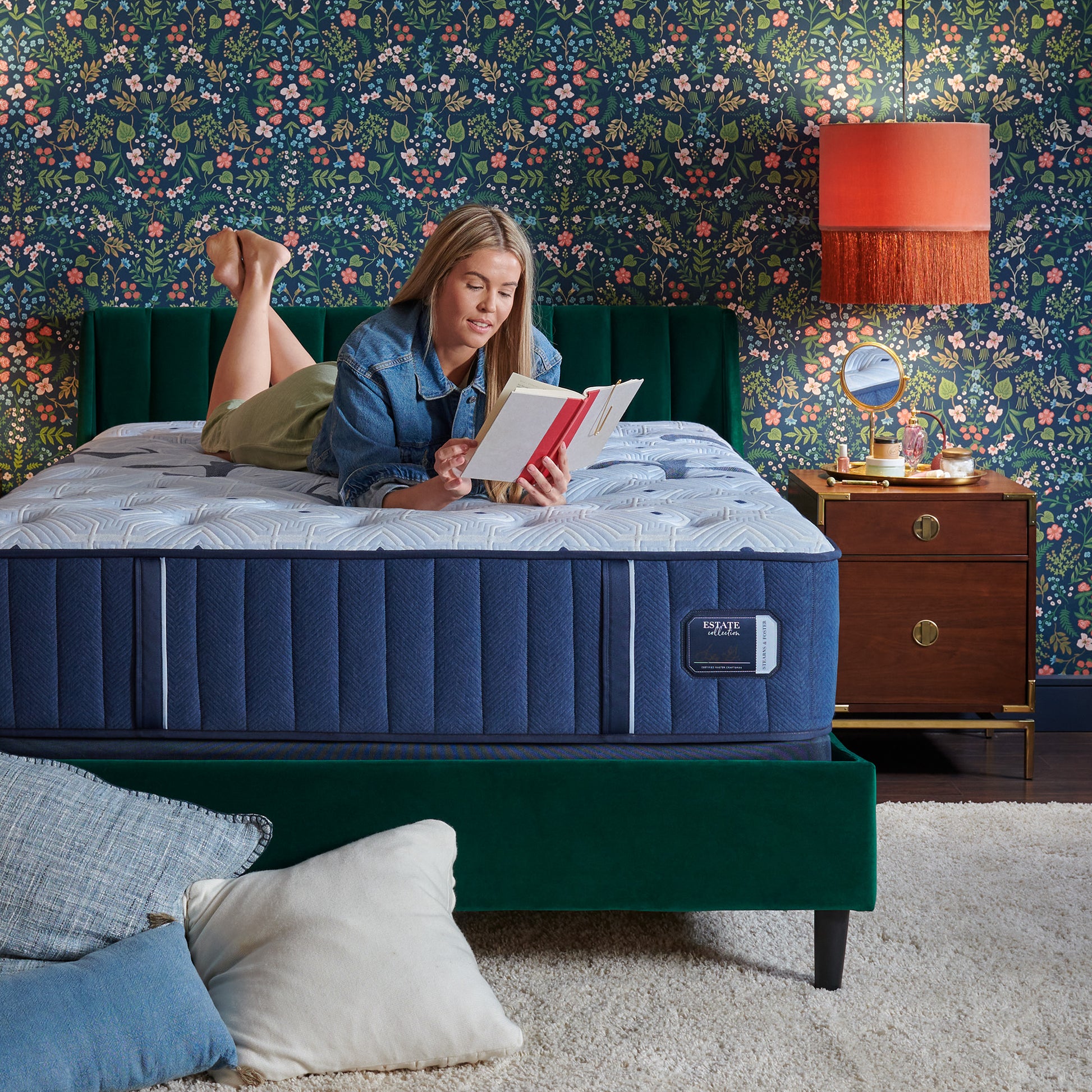 Woman reading a book on a blue mattress in a bedroom with floral wallpaper.
